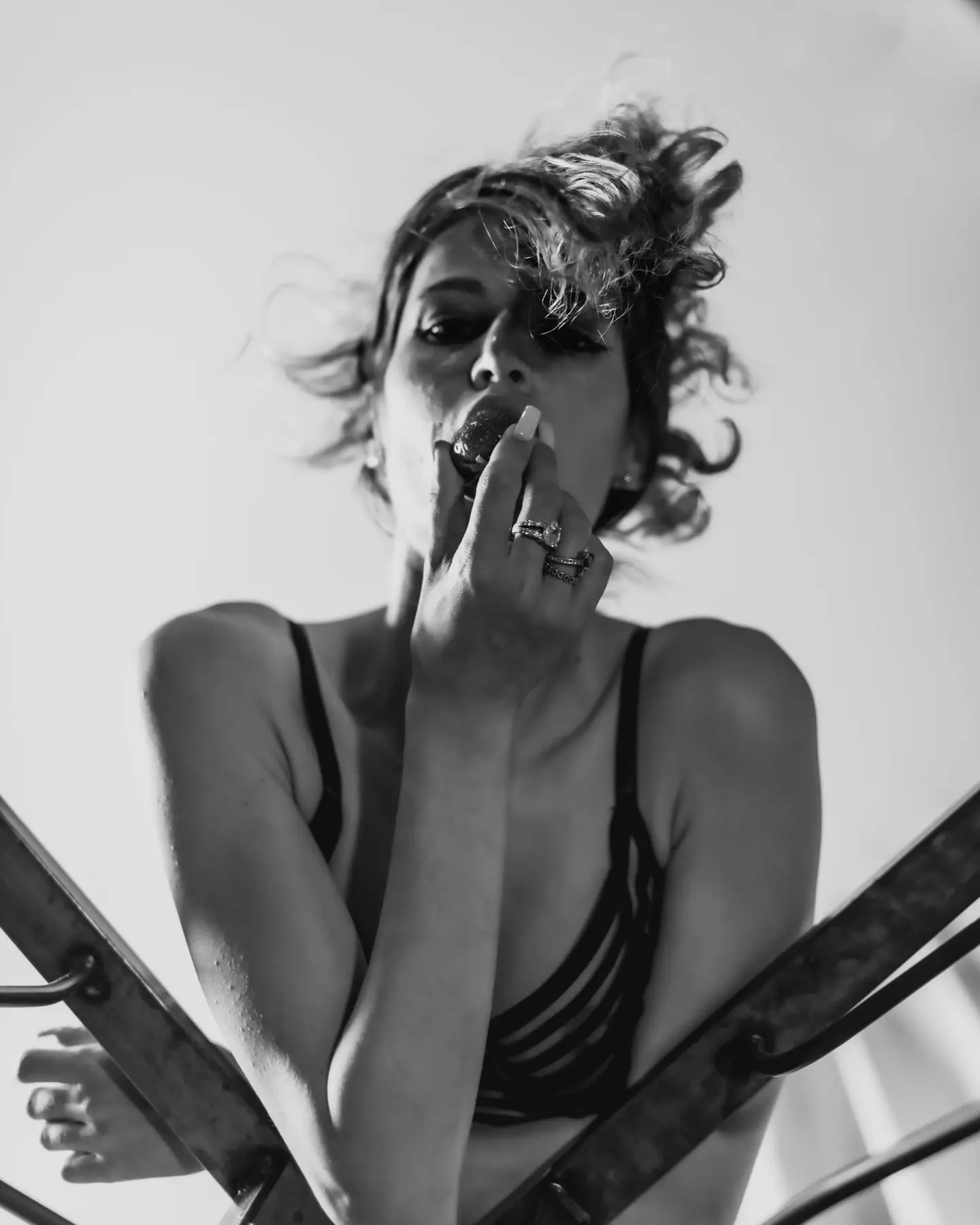Black-and-white intimate portrait shot from a low angle: a curly-haired figure in a dark lace bra brings a strawberry to her mouth, rings catching the light on her hand.