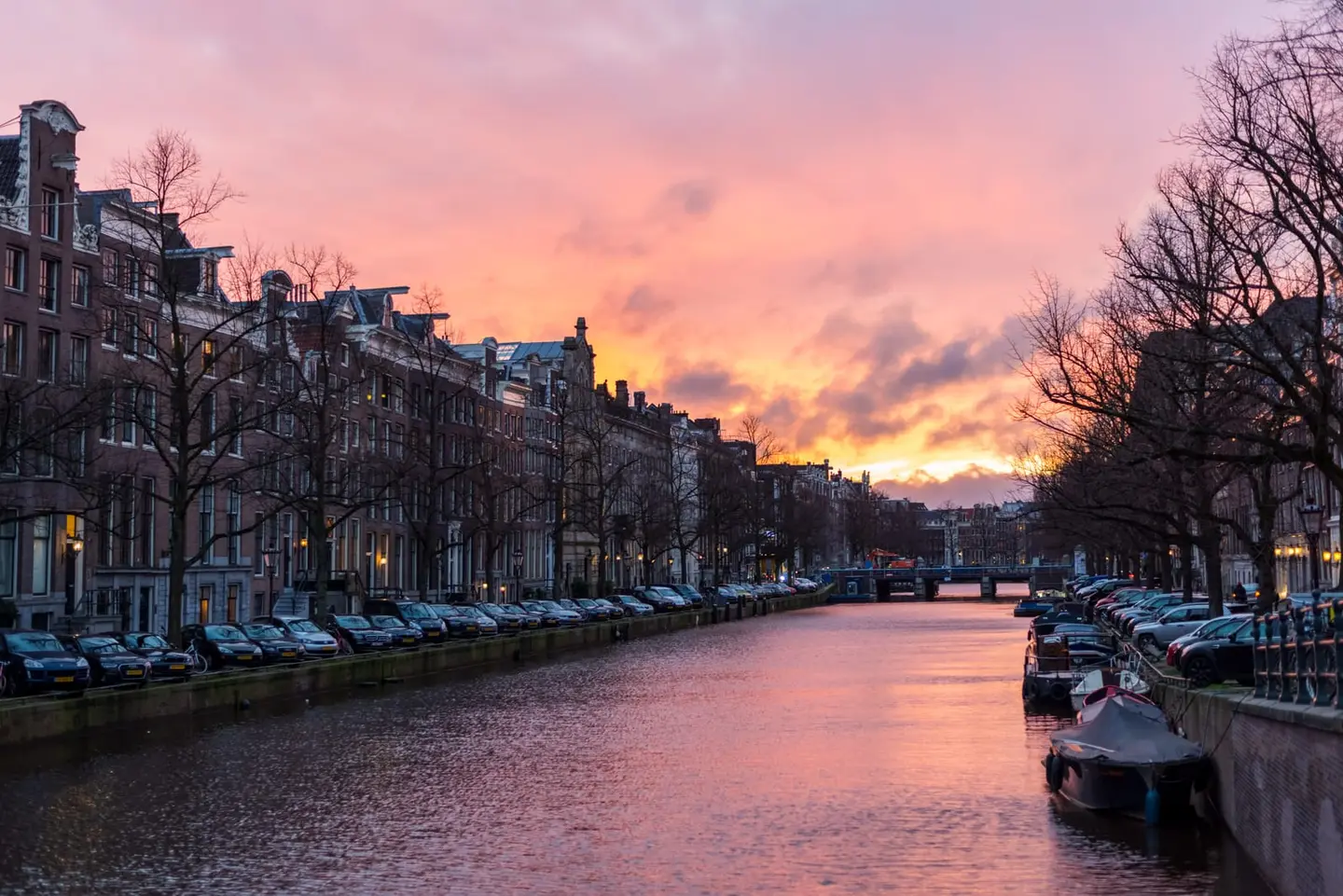 A sweeping sunset of layered rose, amber, and deep violet lights up the sky above a wide Amsterdam canal during the spring 2020 lockdown, the waterway unusually quiet — cars line the quayside but the…