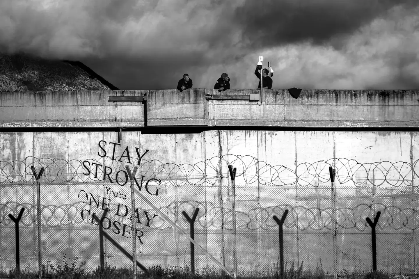 Hand-painted in large block letters on a concrete wall strung with razor wire near Shkoder, northern Albania: STAY STRONG — You're ONE DAY CLOSER.