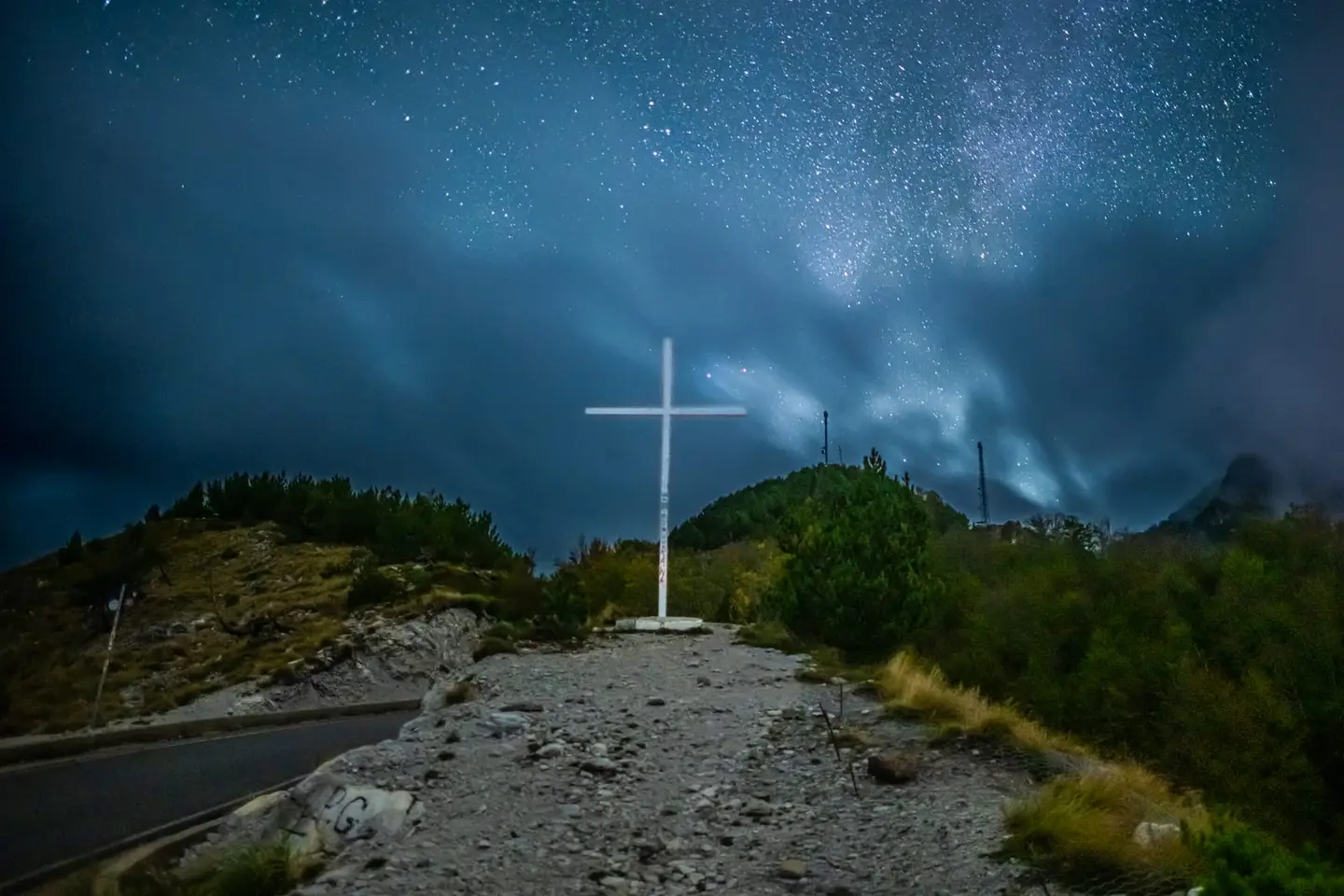 A white painted cross stands at a mountain road junction near Theth in the Albanian Alps under a dramatic night sky.