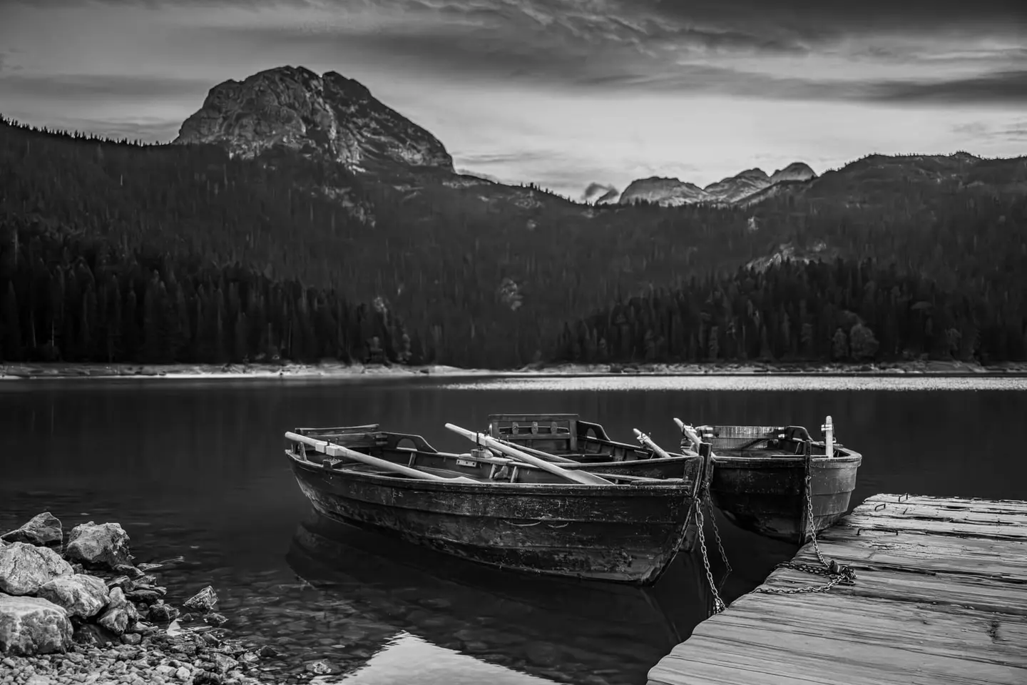 Two weathered wooden rowboats rest chained to a simple plank dock on the still surface of Black Lake (Crno jezero) in Montenegro's Durmitor National Park.