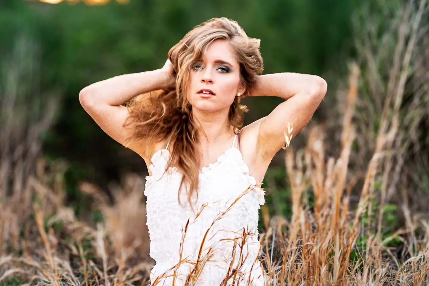 A young woman in a white lace dress raises her hands to her tousled hair amid tall, dry golden prairie grasses — last season's growth still standing against the first green of spring.