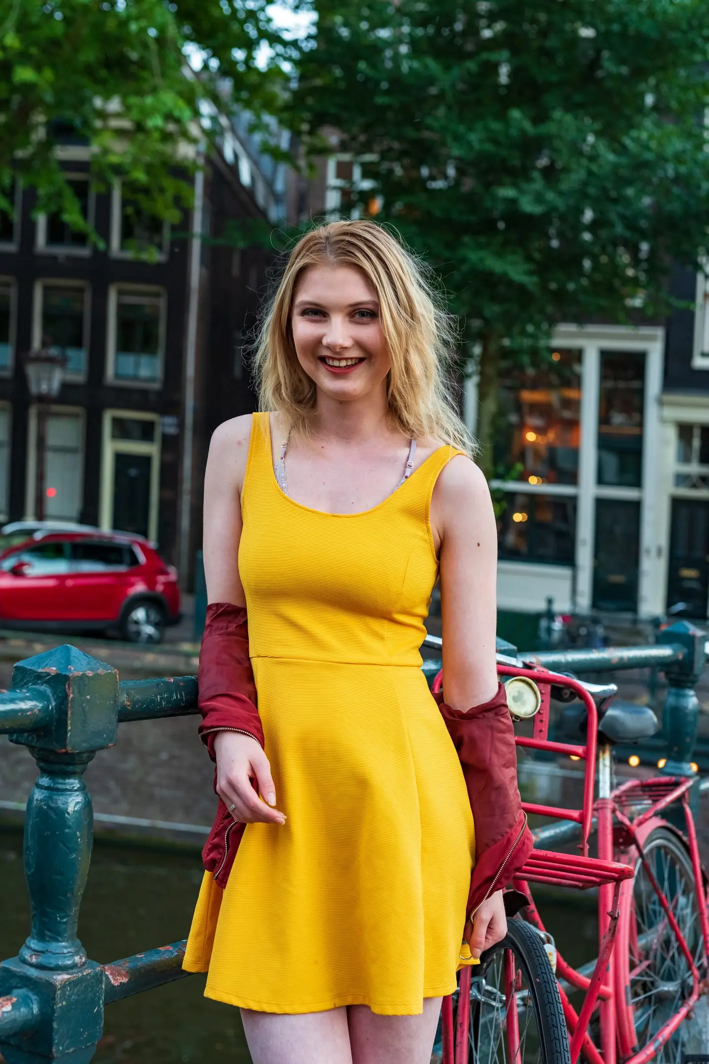 A warm summer portrait on a green-painted canal bridge, leaning on the rail in a bright yellow sundress with a rust jacket tied at the waist.