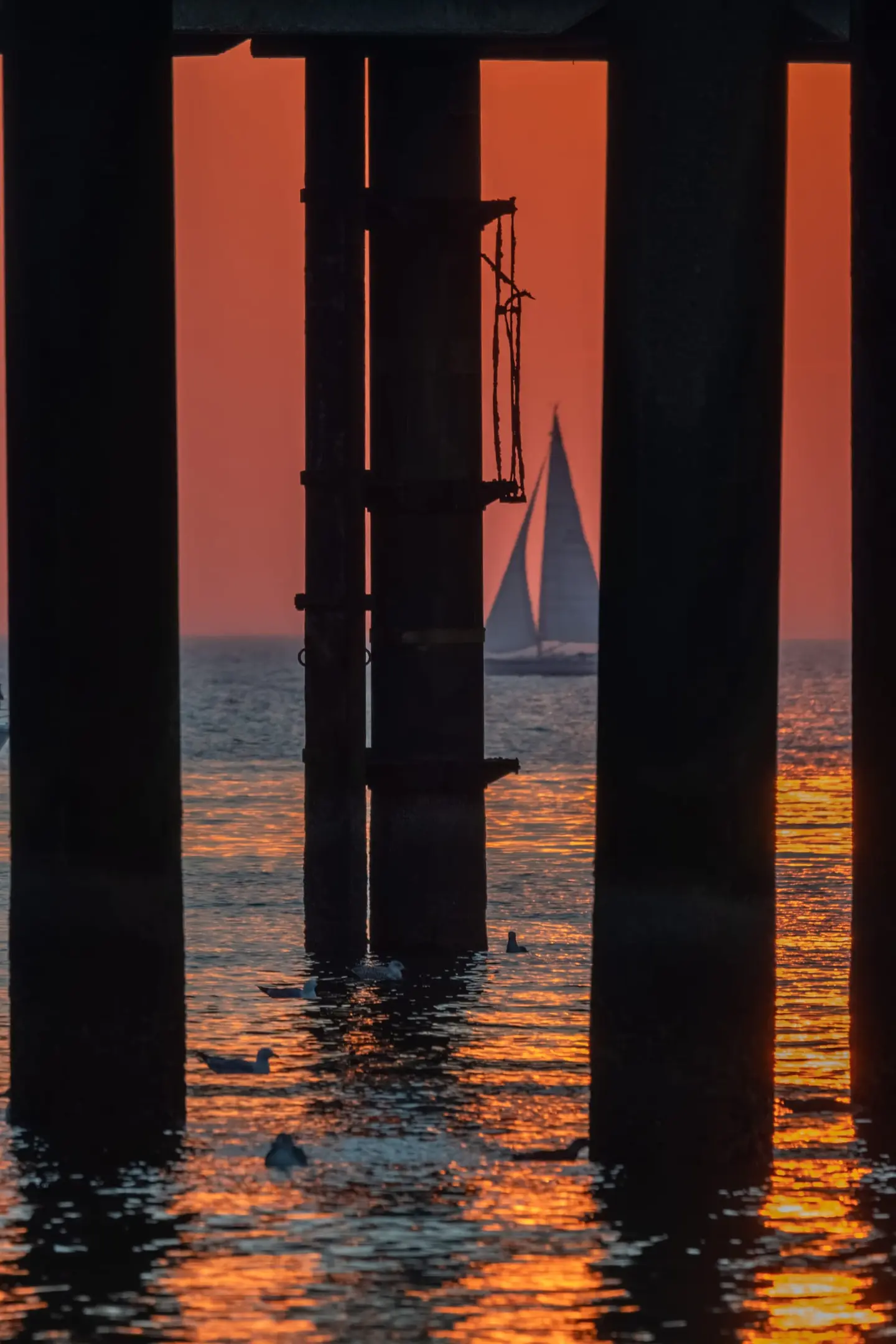 Fiery sunset light pours between the colossal support columns beneath the Scheveningen Pier, where a distant sailboat glides past on the North Sea.