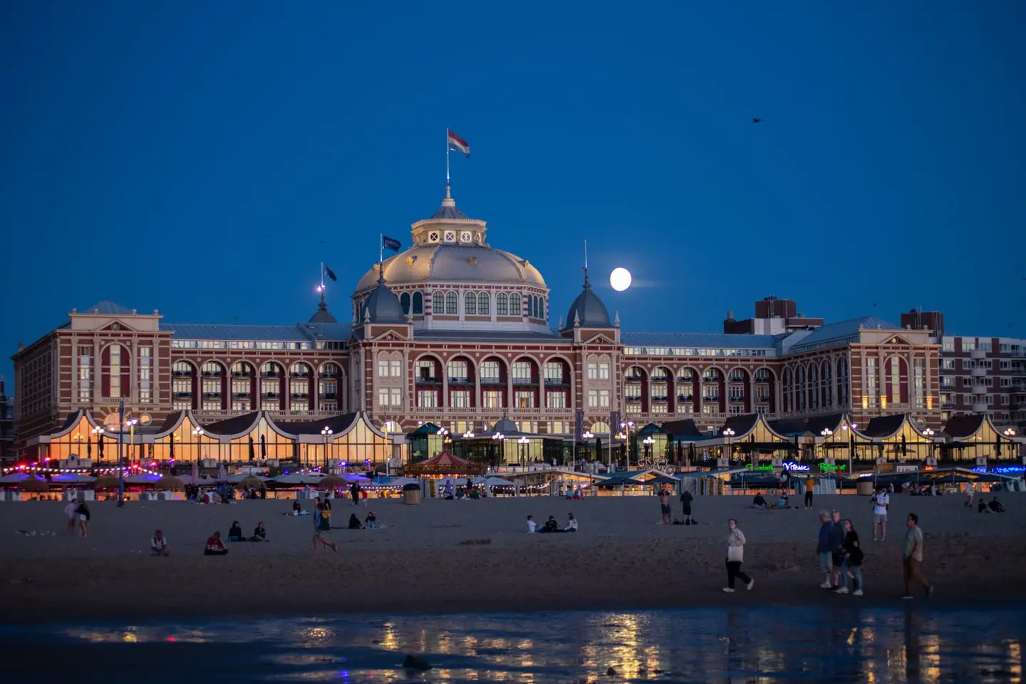 The grand historic Kurhaus of Scheveningen glows warmly beneath a deep blue twilight sky as a full moon rises beside its iconic central dome.