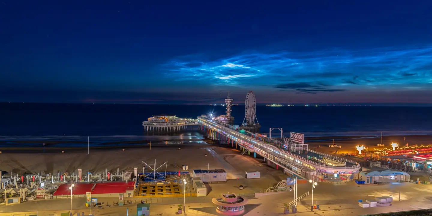 Rare, ethereal blue noctilucent clouds — among the highest and most elusive in the sky — shimmer over the Scheveningen Pier in this sweeping elevated panorama of the Den Haag seafront at deep…