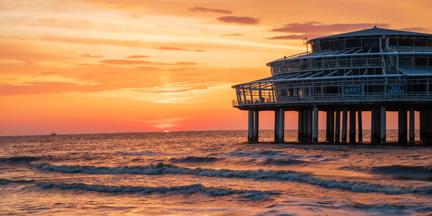 The round glass pavilion of the Scheveningen Pier glows against a blazing orange sky as the sun melts into the North Sea horizon.