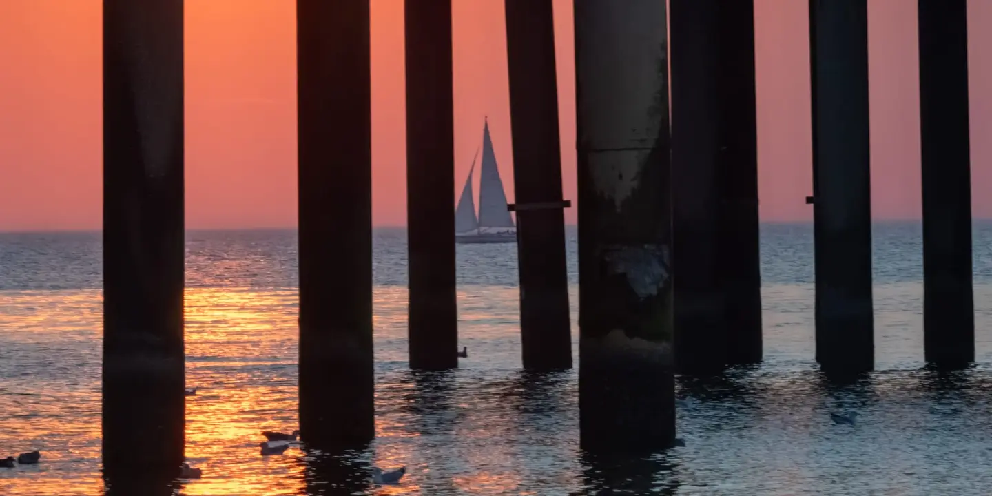 A lone sailboat drifts across the North Sea horizon, framed perfectly between the towering silhouetted pilings of the Scheveningen Pier.