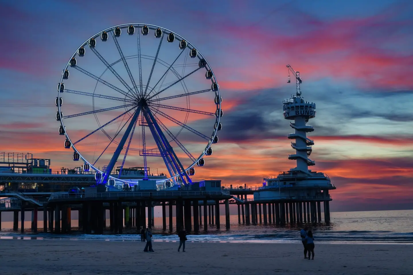 A breathtaking sky of streaked pink, violet, and blue erupts behind the Scheveningen Pier as its Ferris wheel and spiralling bungee tower stand in crisp silhouette.