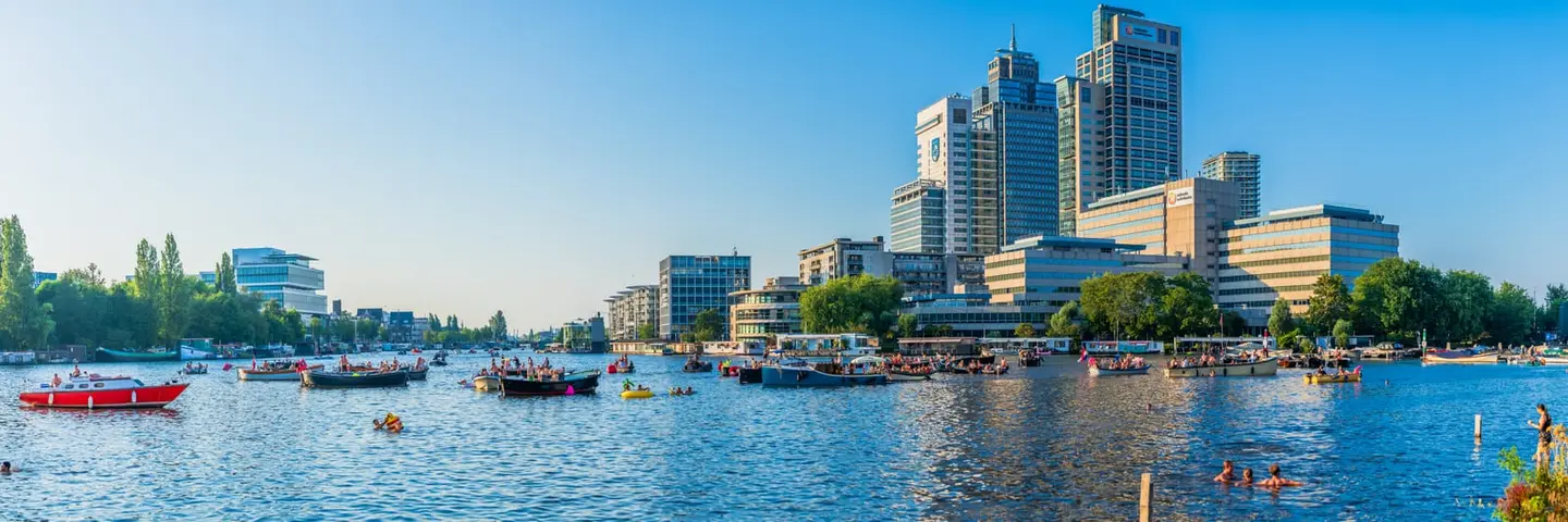 A summer evening on the Amstel in Amsterdam Oost — dozens of small boats, sloops and inflatables drift along the broad river while swimmers cool off at Park Somerlust in the foreground.