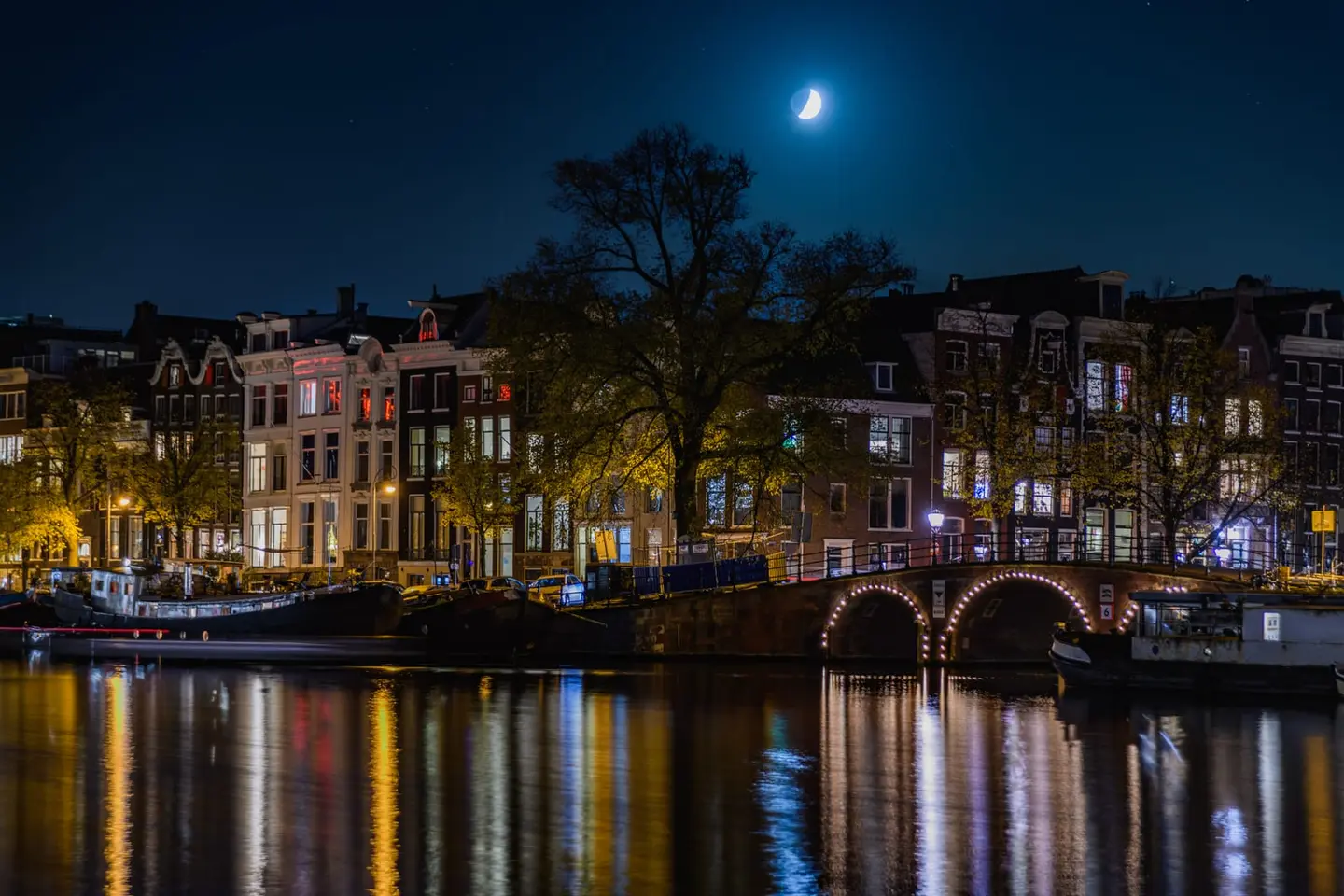 A thin crescent moon hangs in a deep indigo sky above a lit stone bridge on the Amstel from Amsterdam's Weesperbuurt, its arch reflected in perfectly still water below.