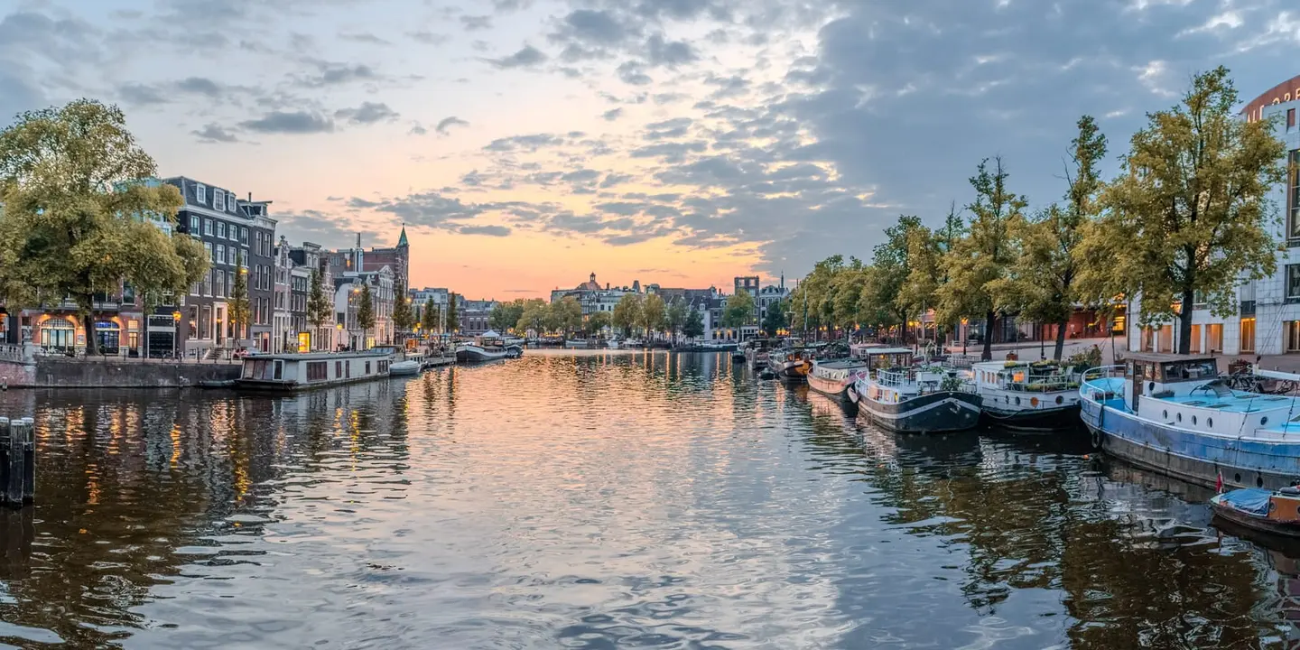 A wide panoramic view takes in the broad expanse of the Amstel as it passes through Amsterdam at sunset, the sky layered in deep rose, amber, and soft grey that reflects across the rippled water…