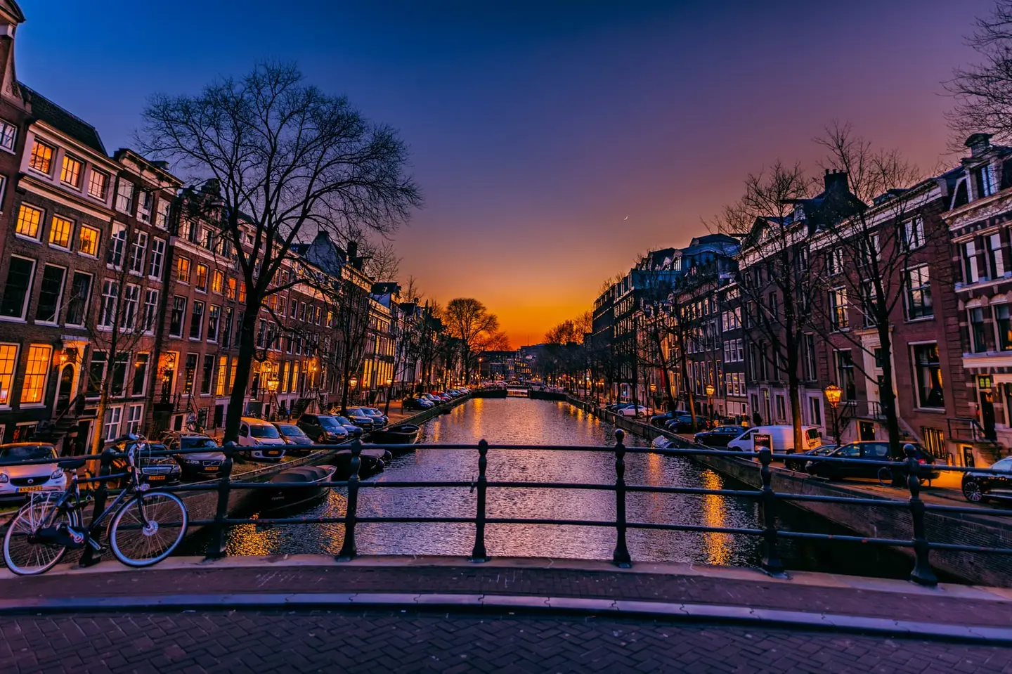 A glowing sunset reflects down an Amsterdam canal lined with classic gabled houses, bare, leafless trees, and a parked bicycle resting against the bridge railing in the foreground.