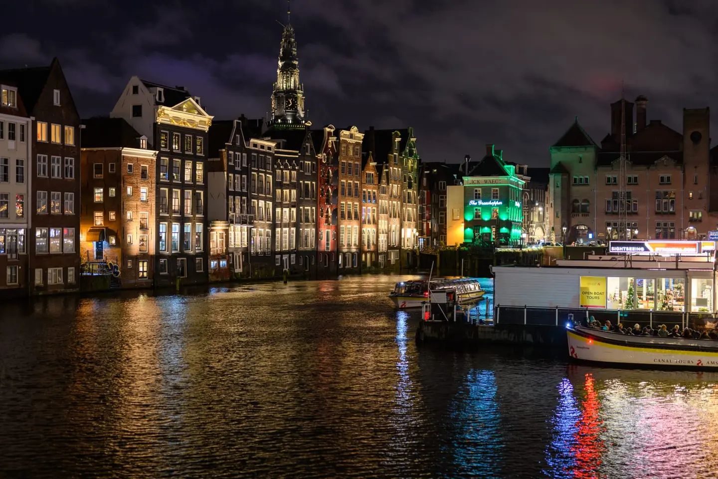 Historic Dutch canal houses glow against a moody night sky while a church spire rises above and colorful reflections shimmer across the still water.