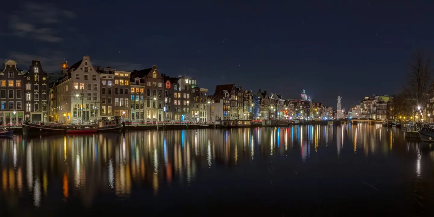 A glass-still Amsterdam canal at night mirrors a long row of historic gabled houses on both banks, with the lit Munttoren rising in the distant centre of the frame.