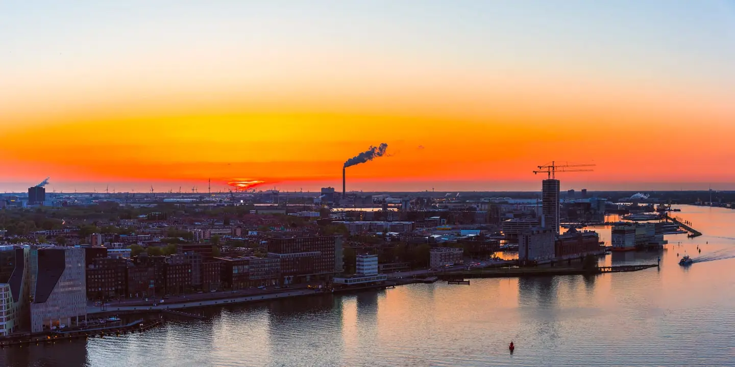 A flat orange sky burns above the IJ waterfront of Amsterdam, with a lone industrial smokestack and a row of harbour cranes silhouetted along the horizon.