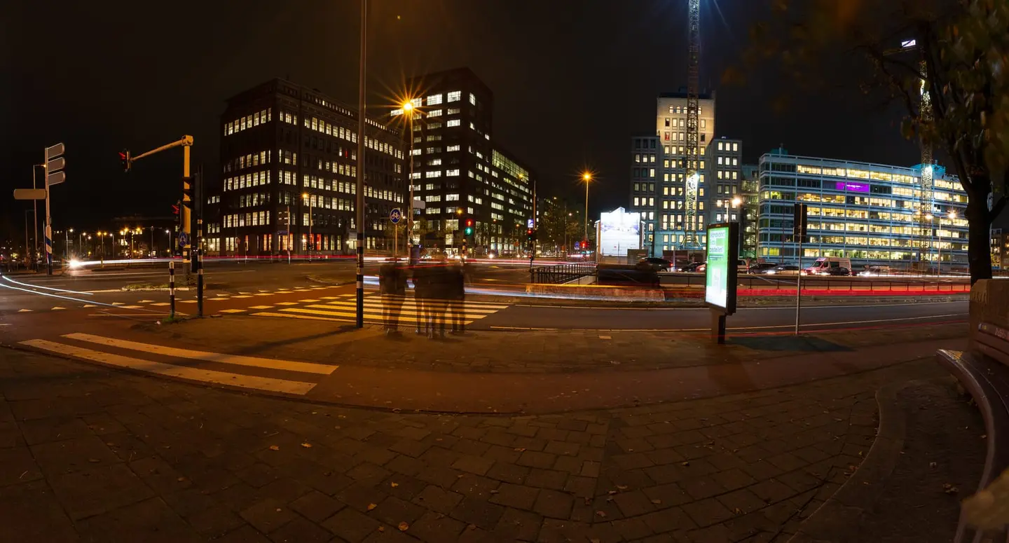 A long-exposure panorama of an Amsterdam city intersection at night — car and tram headlights draw streaks of red and white across the lower frame while ghosted pedestrians blur through the centre…