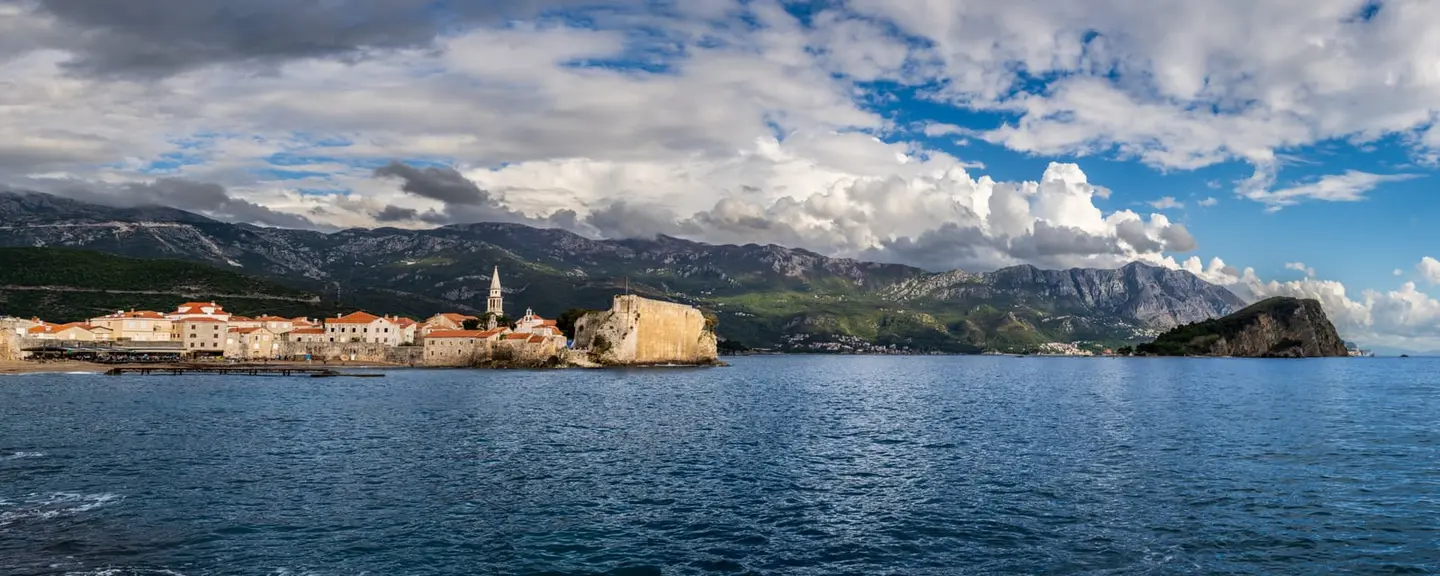 Budva's walled old town juts into the Adriatic on its rocky peninsula, terracotta roofs and the stone campanile catching afternoon light beneath a sweeping bank of cumulus cloud.