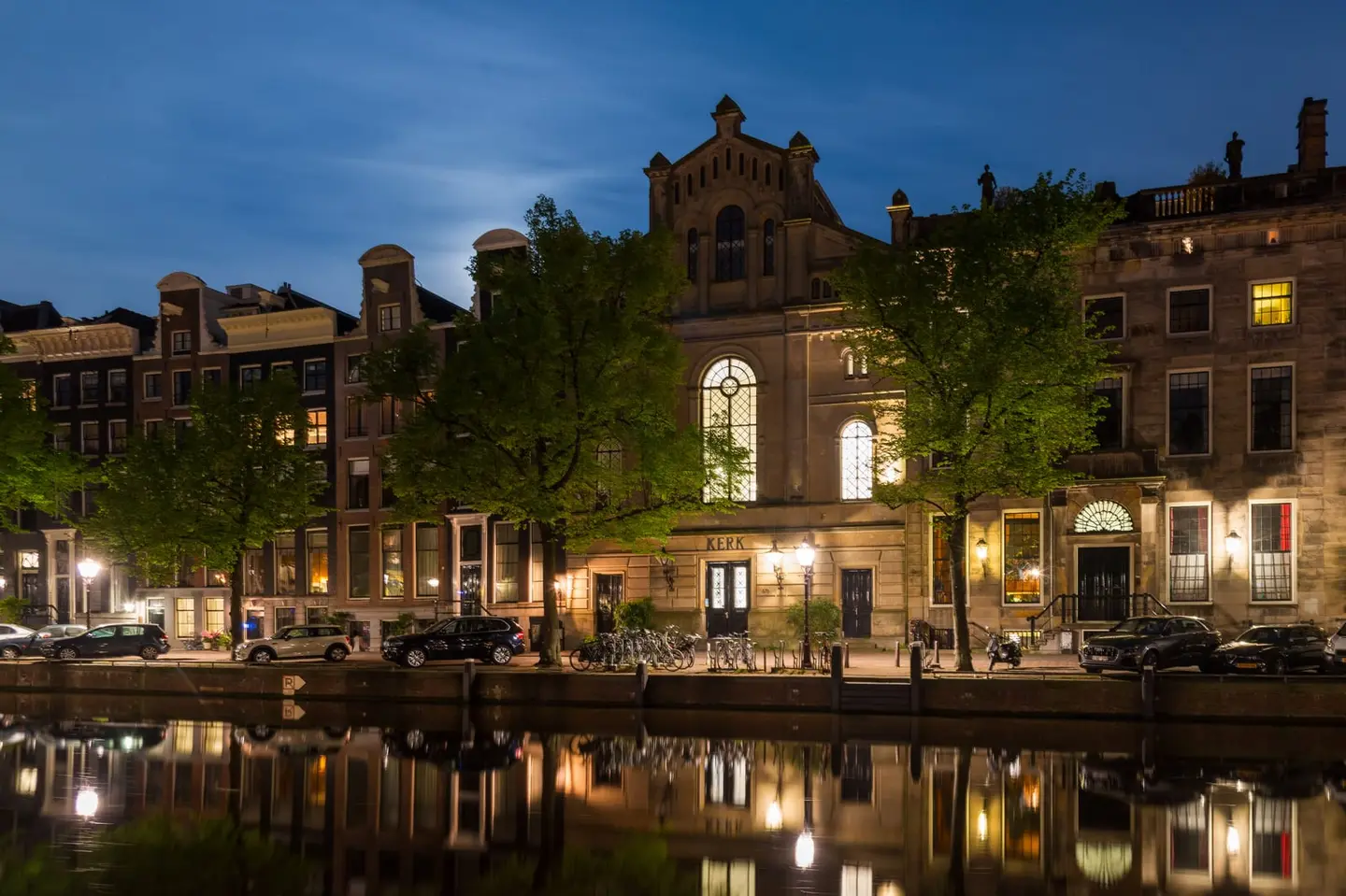 A neo-Renaissance brick church labeled Kerk glows with warm interior light reflected across a still Amsterdam canal in the blue hour of a May evening, the full moon just risen behind the building's…