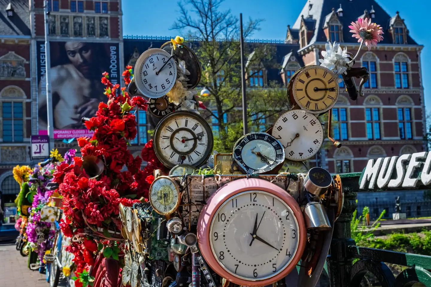 A whimsical art bicycle festooned with vintage clocks and bright fabric flowers stands at Museumplein, the ornate Rijksmuseum facade rising behind it under blue sky.