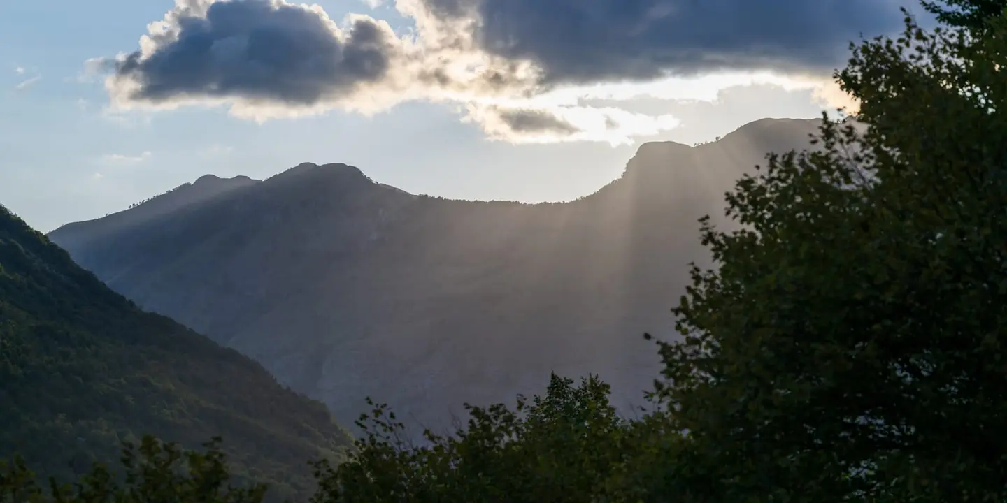Last sunlight breaks behind a long mountain ridge, throwing visible crepuscular rays across the dust and haze of the valley.