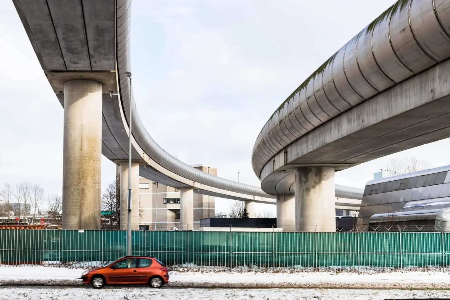Two sweeping concrete metro tracks arc above a roadside fence as a small orange hatchback drives past on a snow-dusted day.