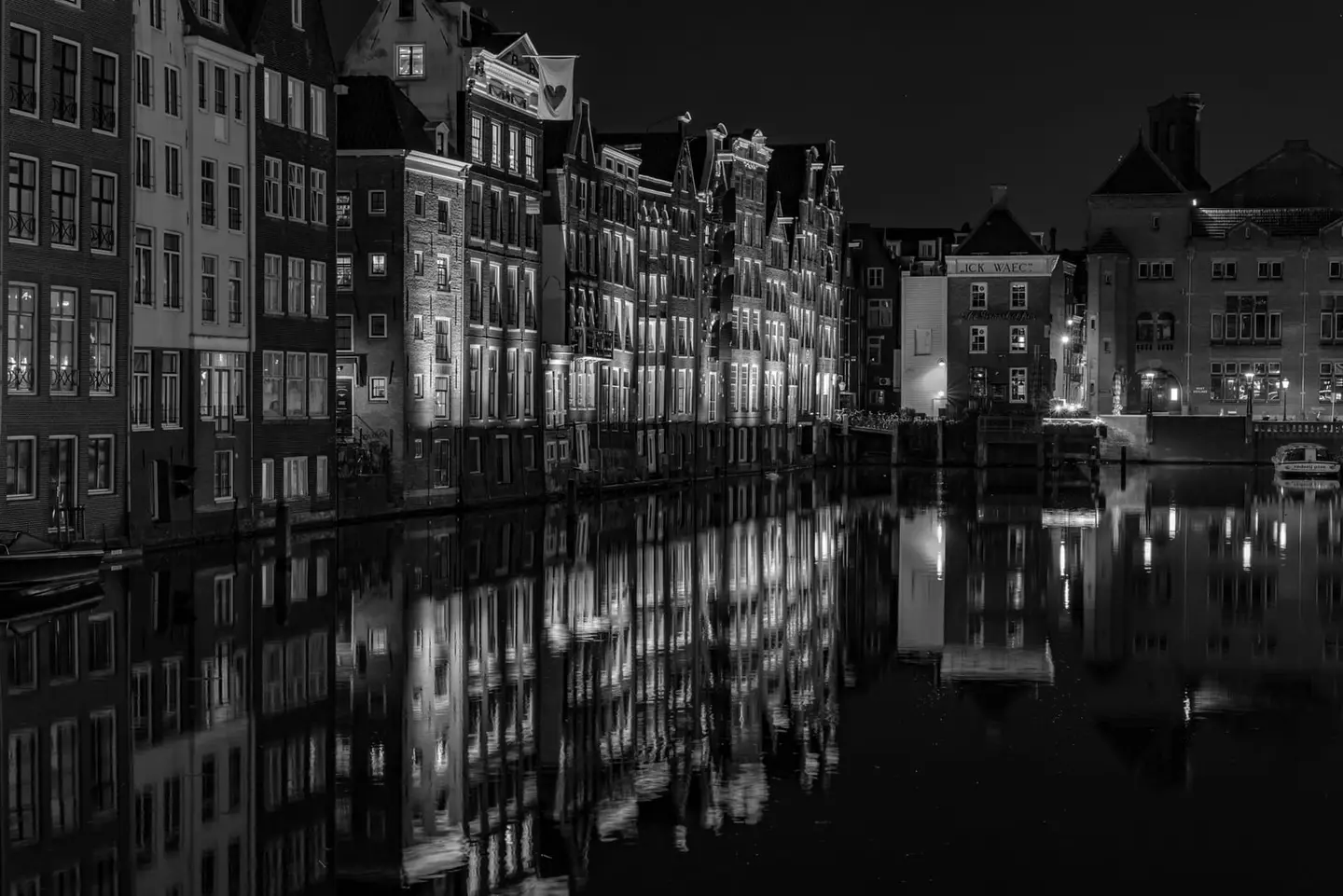 A precisely symmetrical row of Amsterdam's most celebrated canal houses lines the Damrak waterfront in this moody black-and-white night study, their illuminated windows and ornate gable lines…
