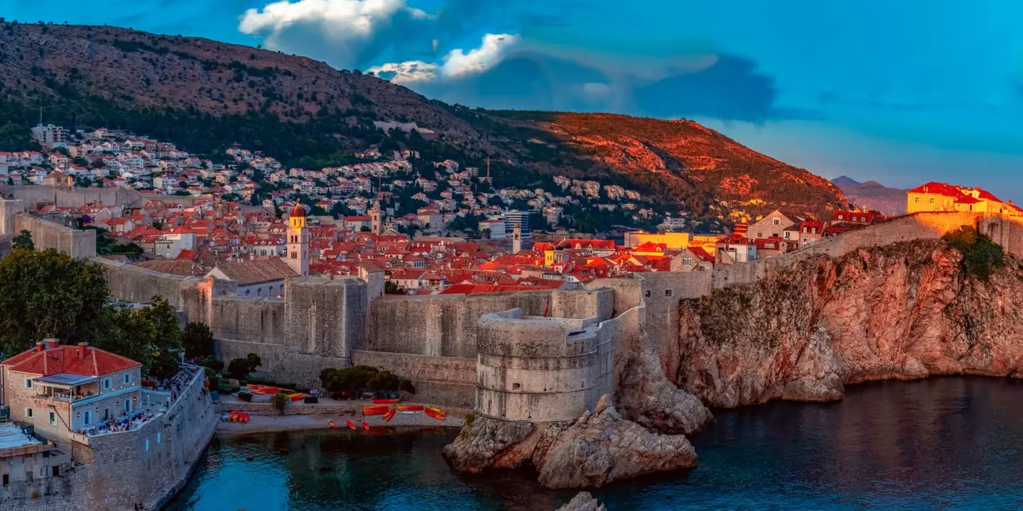 The last light of day washes the medieval ramparts and terracotta rooftops of Dubrovnik old city in warm amber from across the cliff-side bay.