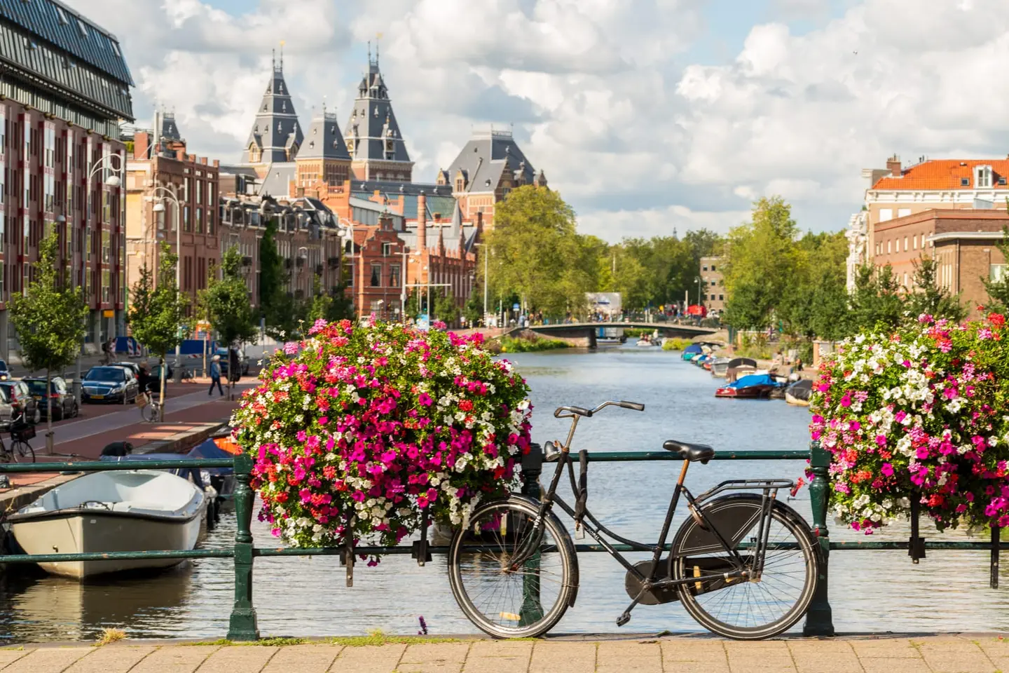 A classic black Dutch bicycle leans against a bridge railing overflowing with deep pink and white petunia flower boxes on an early-autumn afternoon in Amsterdam's Museumkwartier, the bridge framing a…