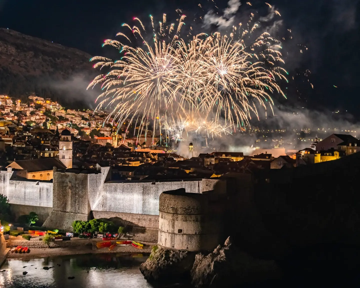Spectacular fireworks erupt over the fortified skyline of Dubrovnik's Old Town, illuminating the medieval city walls and the cylindrical mass of Fort Bokar against the dark Adriatic night.