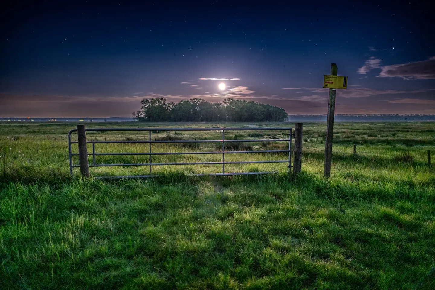 A steel farm gate stands at the edge of De Moerputten nature reserve near Cromvoirt, Noord-Brabant as the full moon rises pale and haloed through thin cloud.
