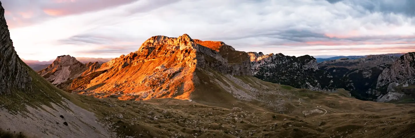 First-light gold rakes across the broad limestone ridge of the Durmitor massif in northern Montenegro, picking out the orange flank of the central peak against deeper-shadowed flanks on either side.