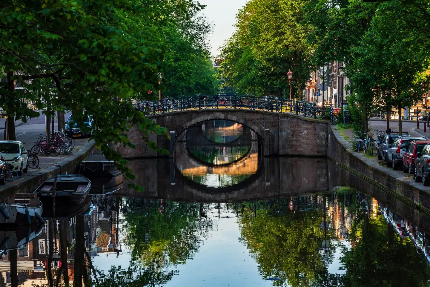 Summer's first light at 6:25am catches a stone arched bridge in the Grachtengordel in perfect bilateral symmetry, the still canal doubling every lamp post, houseboat mooring and leafy canopy.