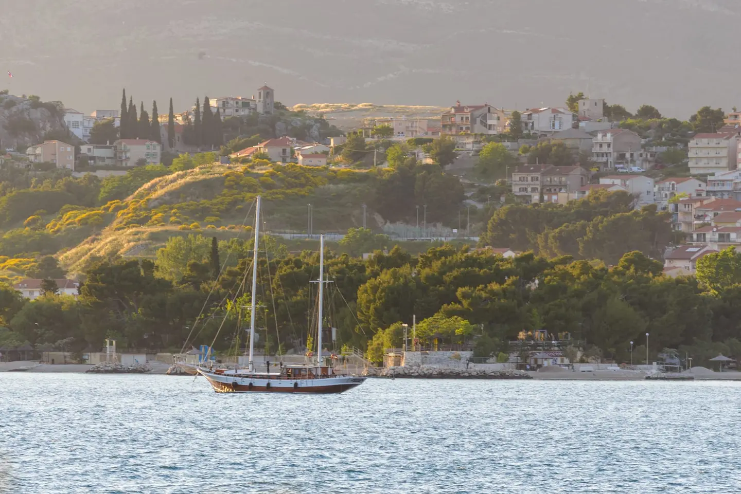 A two-masted wooden gulet rests on calm water below a Mediterranean hillside dotted with red-roofed homes and cypress trees, warmed by golden hour light.
