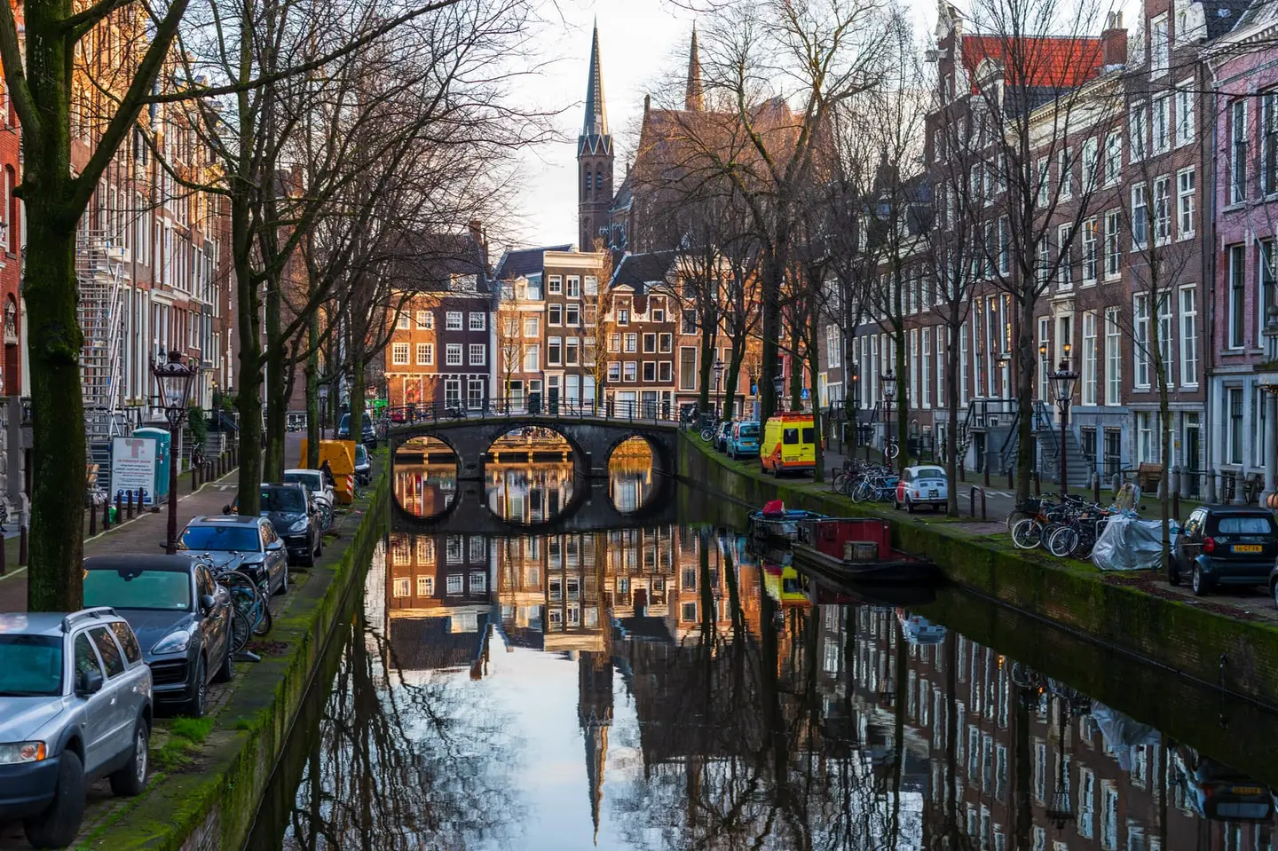 Warm evening light strikes the stone balustrades and arch of a Keizersgracht bridge in the De 9 Straatjes neighbourhood, casting the bridge and canal wall in rich amber while the water mirrors the…