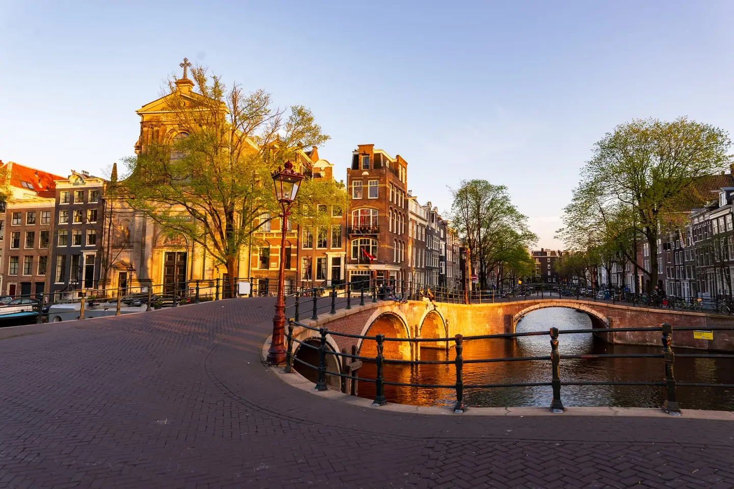 Golden sunset light floods the junction where Keizersgracht meets Leidsegracht in Amsterdam's southern canal ring, basking the stone arch bridges in dreamy gold.