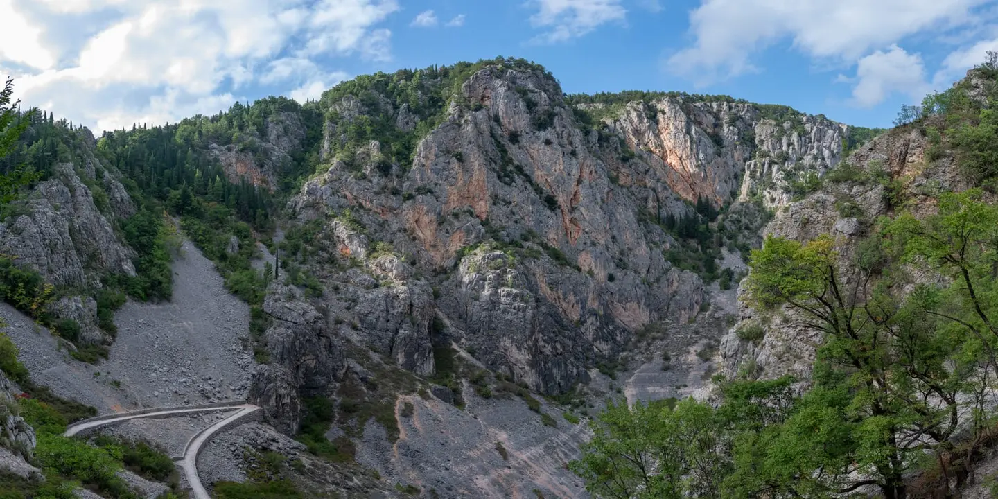 A narrow road bends around the base of a steep limestone gorge, its grey karst walls streaked with orange and stippled with pockets of pine and scrub.