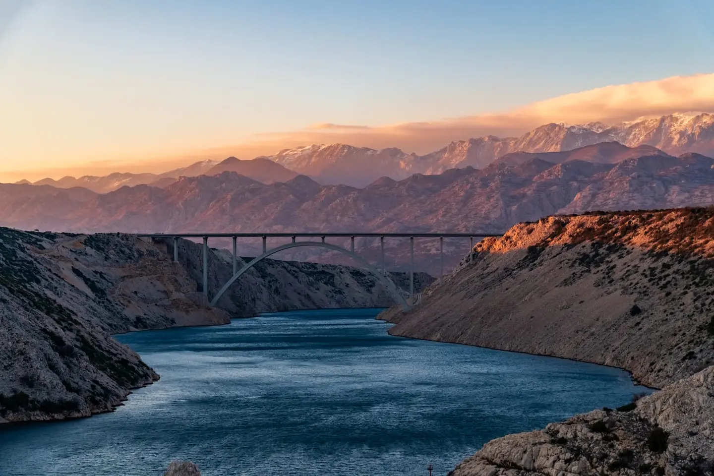 A long arched bridge spans a deep blue canyon inlet as the Velebit mountains glow orange in the last sunlight.