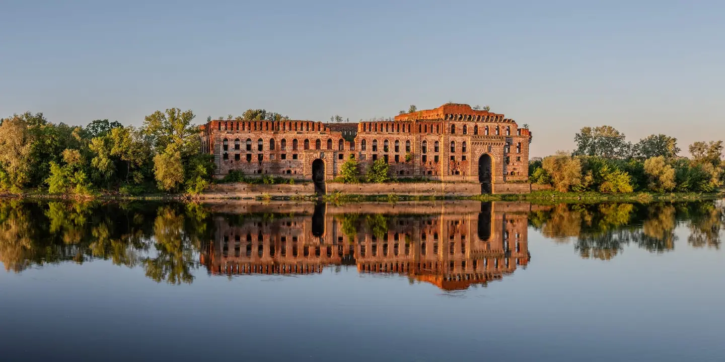 The vast Tsarist-era granary of Modlin Fortress glows in low morning light, its long brick facade and arched gateways mirrored in the still Narew River.