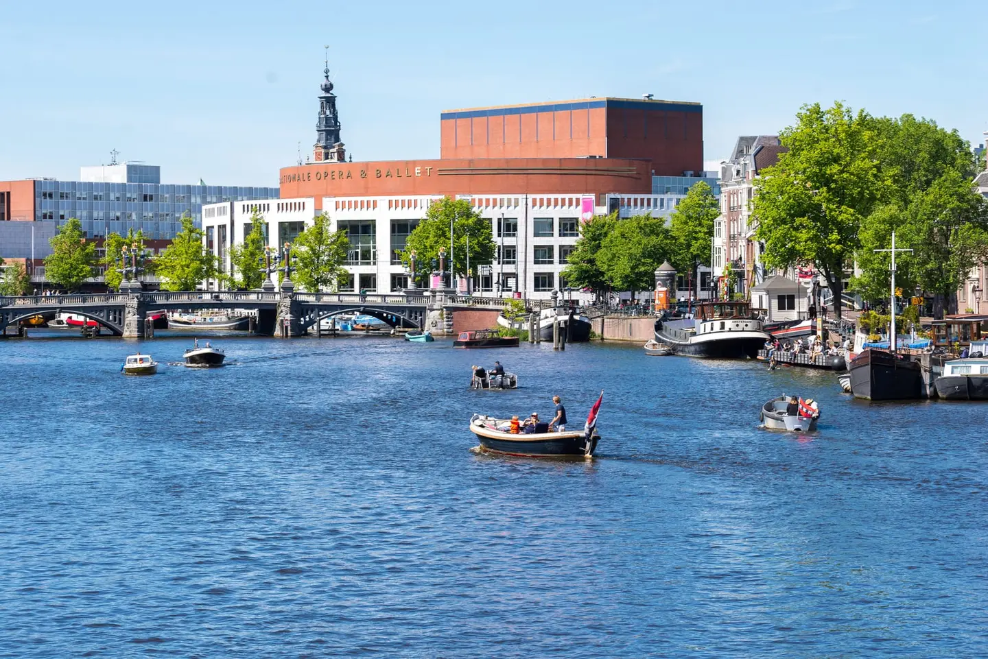 Amsterdam's Nationale Opera & Ballet building — widely known as the Stopera — presents its distinctive brick-and-glass facade across the open water of the Amstel on a bright spring day.