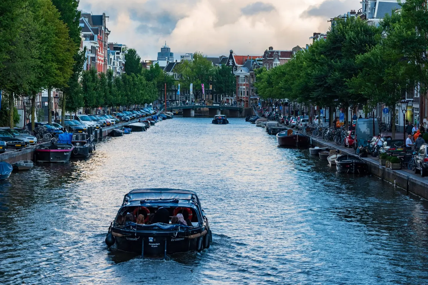 A dark-hulled open pleasure sloop motors away from the camera down a wide Amsterdam canal in the long evening light of early autumn, its wake barely disturbing the reflections of the trees and canal…