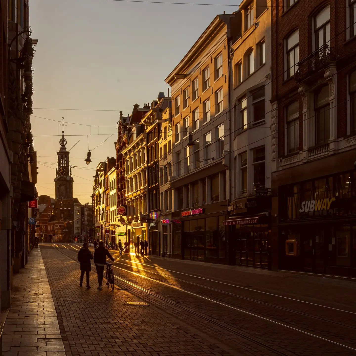 Low golden raking light floods the length of Reguliersbreestraat in Amsterdam at the end of an April day, catching the brick facades and tram tracks in a warm blaze while the shop fronts and…