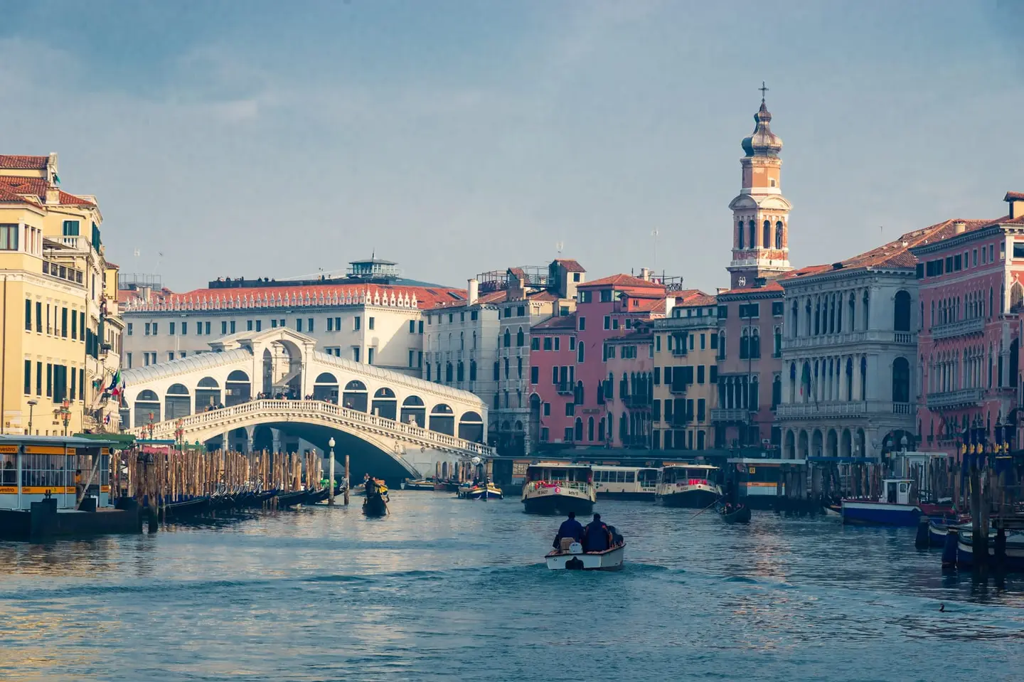 The Rialto Bridge (Ponte di Rialto), Venice's oldest and most celebrated crossing, spans the Grand Canal in warm afternoon light, its white Istrian stone arches rising above gondoliers and water…