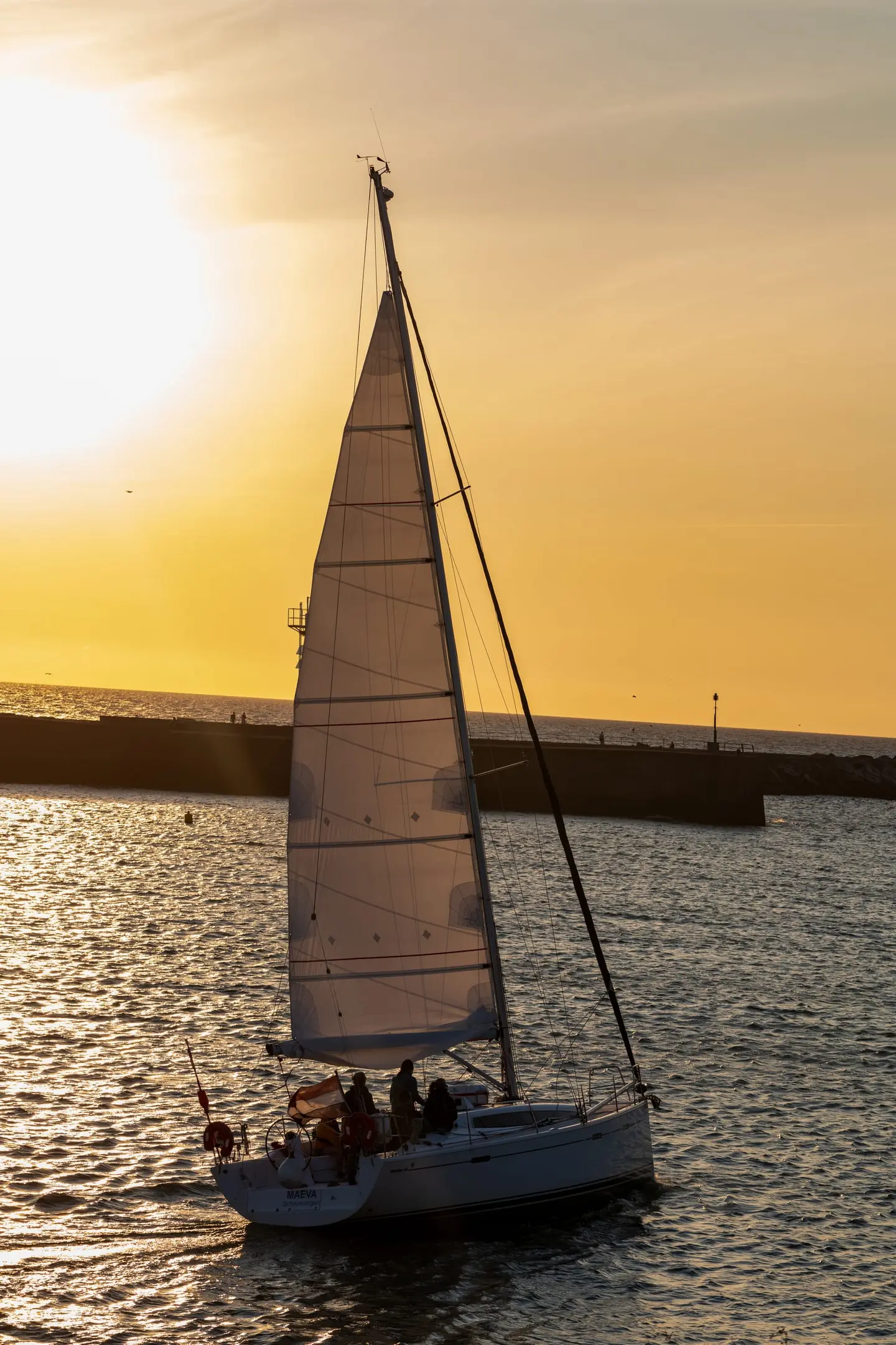 A lone sailboat glides past a stone breakwater toward Scheveningen harbor as the low sun glows through its white sail, casting amber light across calm water and a soft golden sky.