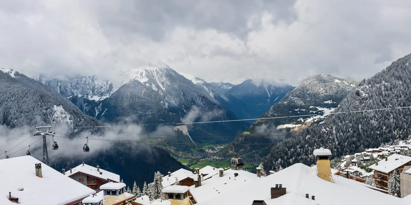 Fresh snow covers the chalet rooftops of a high-altitude ski village while two cable car lines climb out of the frame toward cloud-wrapped peaks.