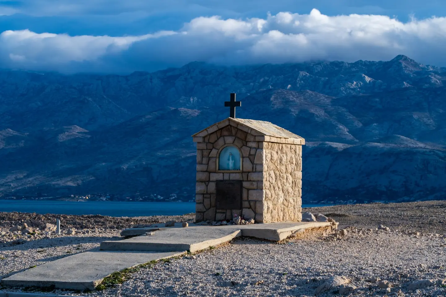 A small stone wayside shrine stands alone on a windswept karst plateau, its carved niche sheltering a blue-robed Madonna figure beneath a simple cross.