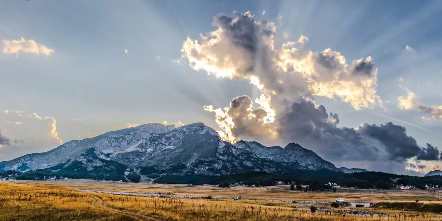 Late-afternoon sunbeams pour through a break in heavy storm clouds, lighting the upper slopes of a long mountain ridge while a small red-roofed town settles into the wide grass valley below.