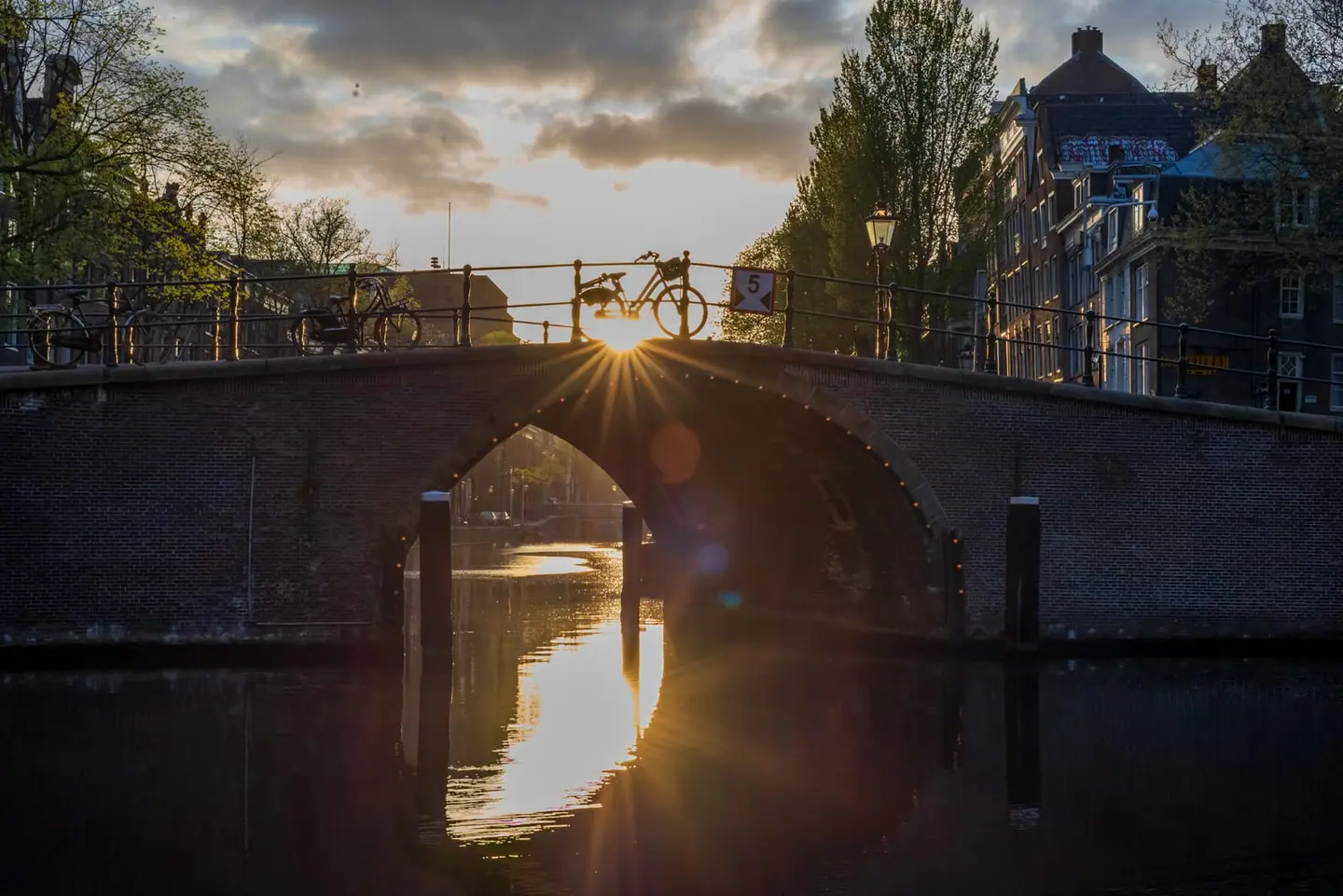 The setting sun bursts through a parked bicycle on an arched brick canal bridge, casting a golden starburst and shimmering reflection along the still water of the grachtengordel.