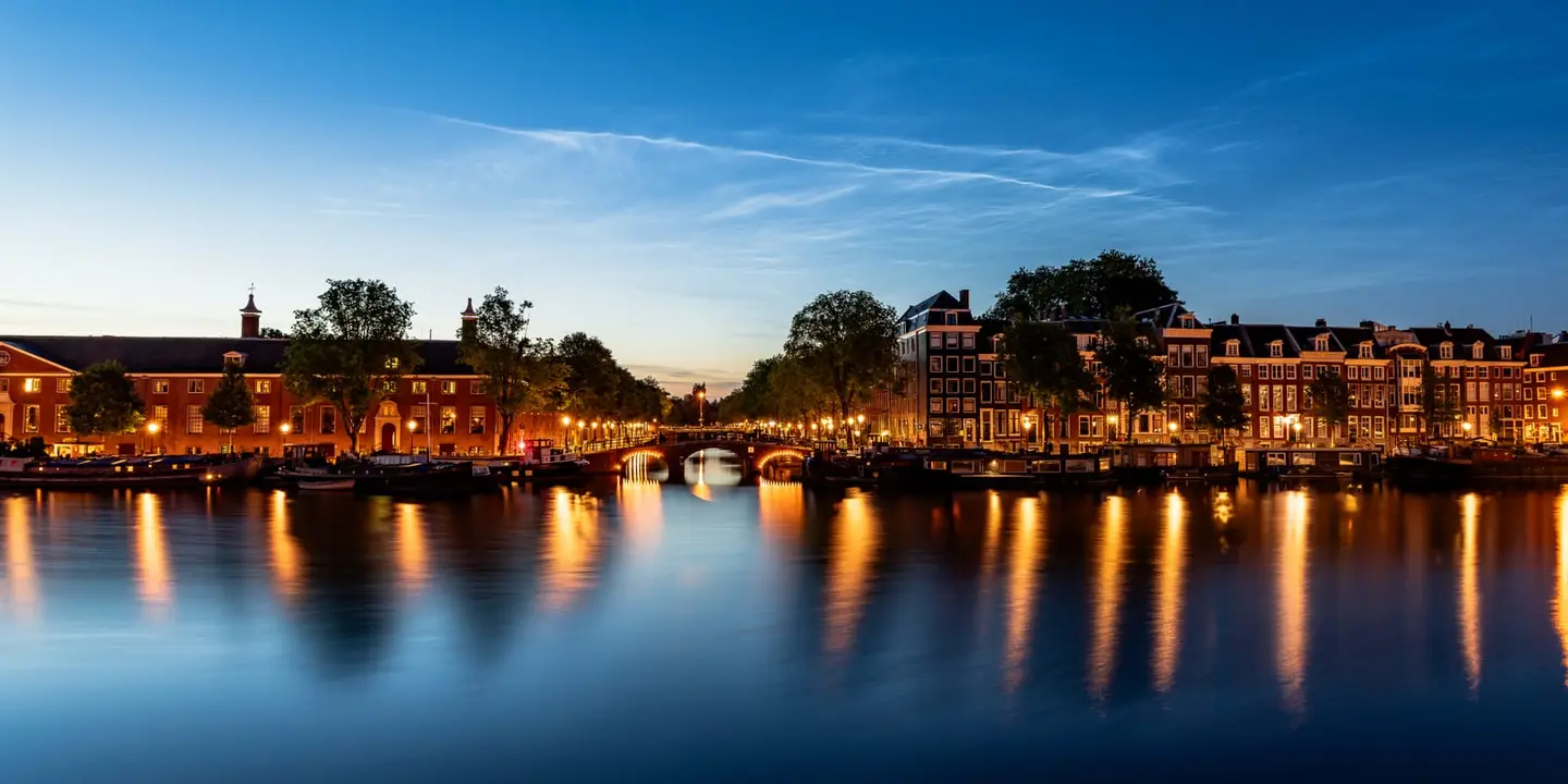 Rare noctilucent clouds — the highest clouds in the atmosphere, lit from beyond the horizon and visible only in deep midsummer twilight — shimmer in electric-blue wisps over the river Amstel in…