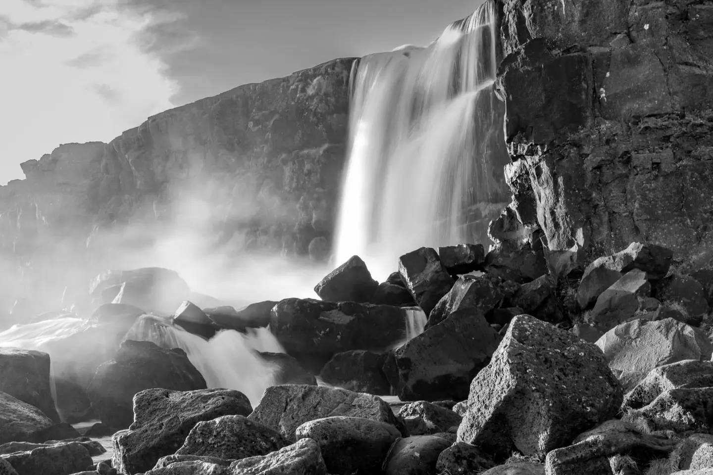 A long-exposure study of a waterfall pouring over a wall of columnar basalt into a field of tumbled volcanic boulders in Iceland.